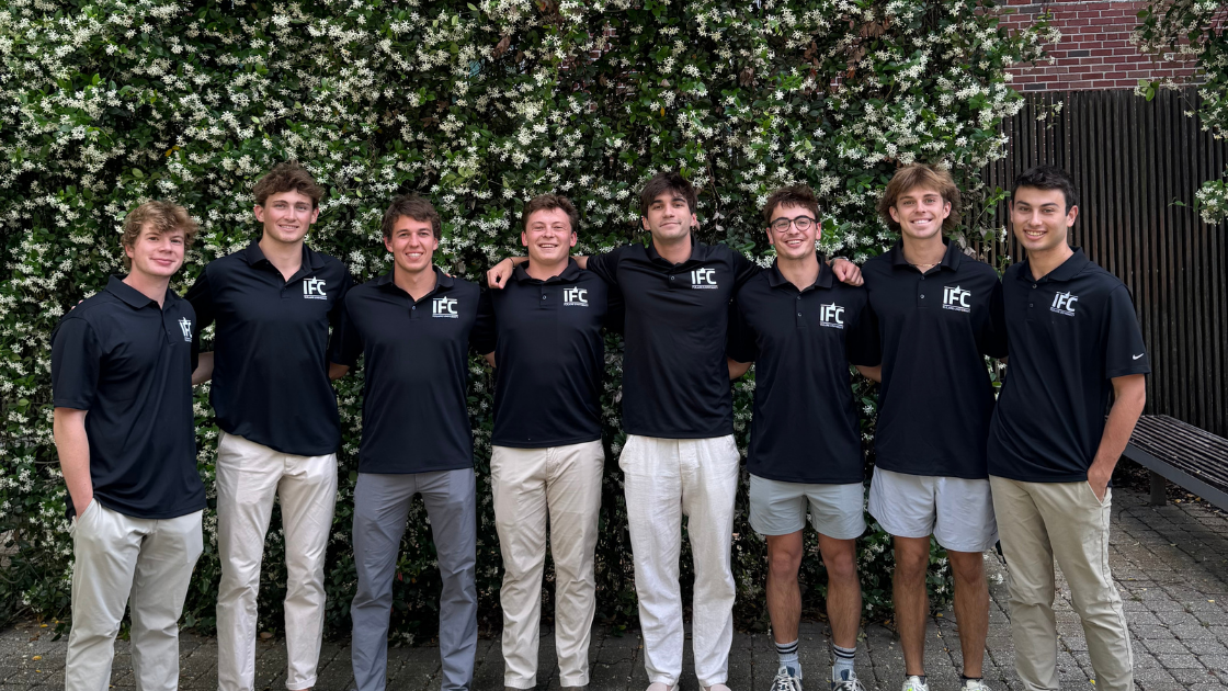 Eight young men in uniform navy polo shirts and light pants, arms around each other, by an ivy wall.
